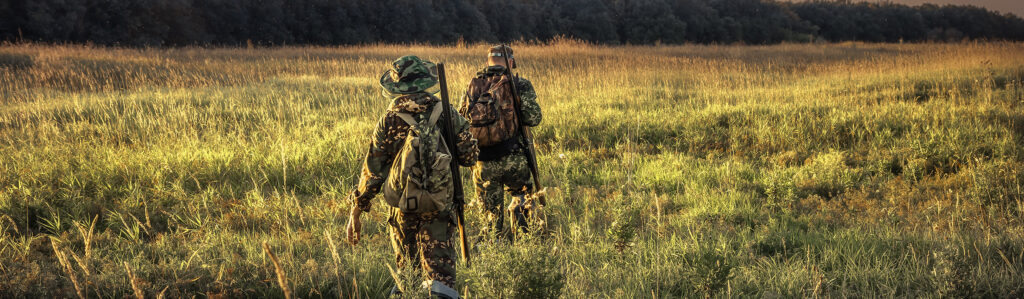 Father and son hike through a large field with their rifles slung over their shoulders while hunting.