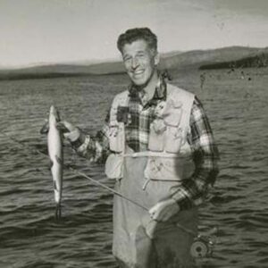 A black and white photograph of Lee Wulff standing knee-deep in a lake smiling at the camera while holding a fish in his hand.