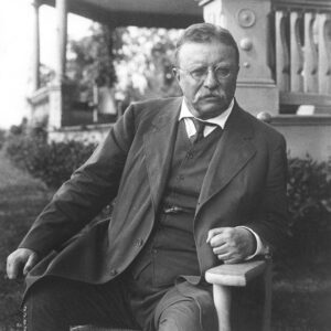 A black and white photograph of Theodore Roosevelt sitting in a chair on the porch of a house.