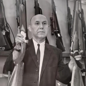 Ira/Irv Joffe stands behind his gun counter holding two rifles and looking off camera to an interviewer.
