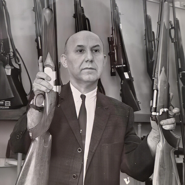 Ira/Irv Joffe stands behind his gun counter holding two rifles and looking off camera to an interviewer.