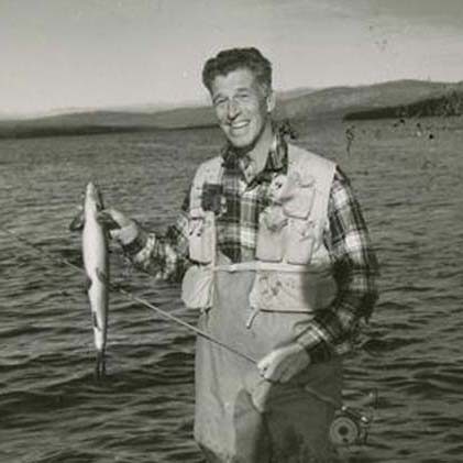 A black and white photograph of Lee Wulff standing knee-deep in a lake smiling at the camera while holding a fish in his hand.